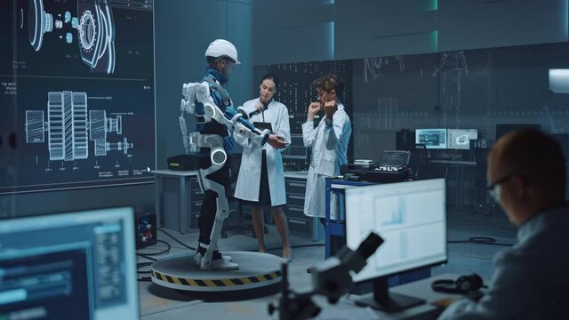 In Robotics Development Laboratory: Engineers and Scientists Work on a Bionics Exoskeleton Prototype with Person Testing it. Designing Wearable Exosuit to Help Disabled People, Warehouse Workers
