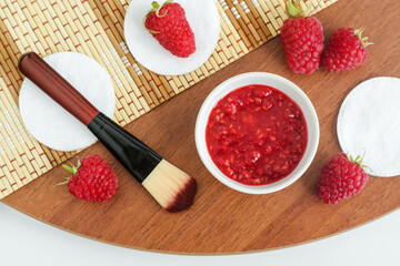 Diy raspberry mask in a small white bowl, cosmetic brush and cotton pads. Homemade beauty treatment and spa recipe. Top view, copy space. 