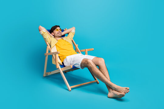 Full length profile side photo of positive guy traveler relax rest beach sun bathing lie comfort deckchair stretch hands wear white shorts isolated over blue color background