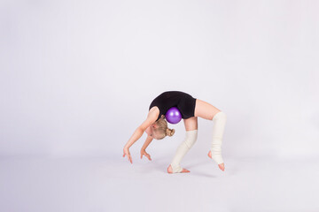a girl gymnast in a black swimsuit does a bridge exercise with a ball on a white isolated...
