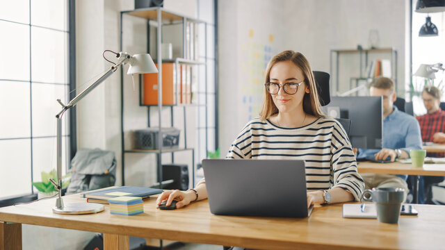 Beautiful Young Woman in Glasses is Working on a Laptop in a Creative Business Agency. They Work in Loft Office. Diverse People Working in the Background. She's in a Good Friendly Mood. - Powered by Adobe