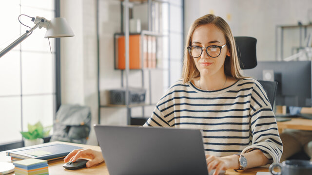Beautiful Young Woman in Glasses is Working on a Laptop in a Creative Business Agency. They Work in Loft Office. Diverse People Working in the Background. She's in a Good Friendly Mood.