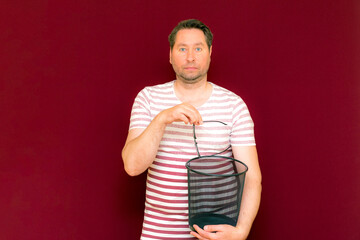weird middle age man wears striped t shirt throws glasses in the trash bin on a burgundy background.