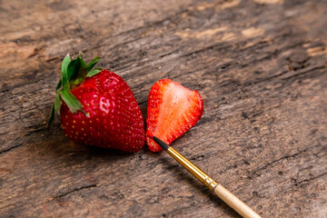 One whole strawberry and one half strawberry with paint brush on a dark wooden surface with space to write on the right.