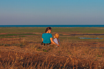 Mother and daughter sitting by lakes on a nature lanscape. A little girl with long blond hair braid hugging with mom outdoor. Loving child embrace her mommy. Mother's Day family greeting card faceless