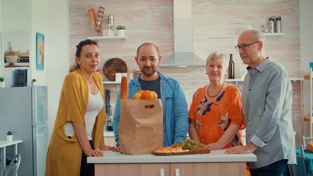 Video portrait of happy extended family smiling at the camera, sitting in the kitchen. People in dining room around the papper bag with groceries looking at the web cam