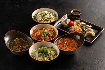 Symmetrical composition of an-Asian and Pan-Asian cuisine on a stylish black concrete kitchen table. Dumplings, soups, side dishes of rice, egg noodles, glass noodles and vegetables.