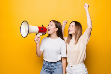 Two irritated young women girls friends scream in megaphone isolated on yellow background . People...