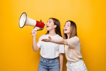 Two irritated young women girls friends scream in megaphone isolated on yellow background . People lifestyle concept.