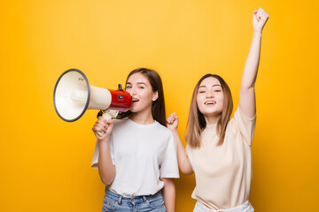 Two irritated young women girls friends scream in megaphone isolated on yellow background . People...