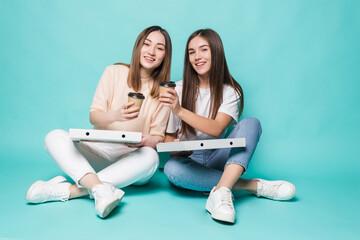 Two women friends sitting on the floor drink coffee to go and eat pizza isolated on turquoise...