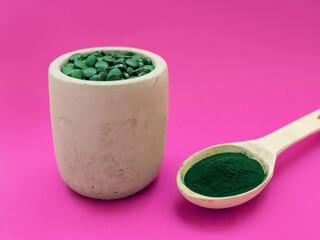 Green hawaiian spirulina in tablespoons pills and powder on pink  background. Super food, healthy lifestyle, healthy supplements concept