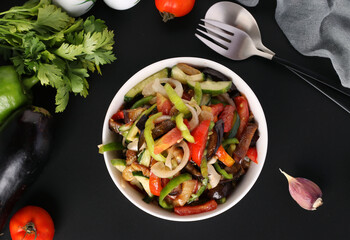 Salad with eggplants, tomatoes, peppers and cucumbers in white bowl on black background, Top view, Closeup