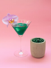Liquid spirulina green drink with Phalaenopsis in cocktail glass and spirulina pills on pink background. Super food, healthy lifestyle, healthy supplements concept