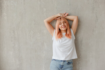 Young joyful woman in white t-shirt with dyed hair posing at wall with raised hands