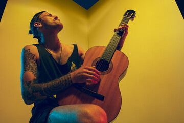 Obraz premium Young musician with guitar performing on yellow background in neon light. Concept of music, hobby, festival, entertainment, emotions. Joyful, inspired guitarist. Colorful portrait of artist.