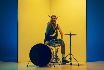 Rockstar. Young musician with drums performing on yellow background in neon light. Concept of...