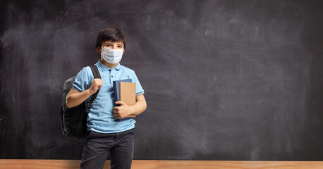 Boy in school with a backpack and books standing in front of a school blackboard and wearing a face mask