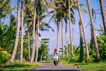 Surfer girl in swim suit rides motorbike on road to the ocean with palm trees in sunny day with blue sky