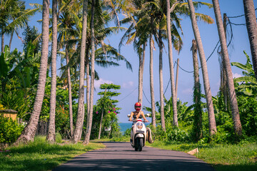 Surfer girl in swim suit rides motorbike on road to the ocean with palm trees in sunny day with blue sky