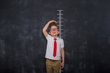Little kid boy measuring himself. School boy measuring his growth in height against a blackboard scale. Back to school. Pupil in uniform - obrazy, fototapety, plakaty