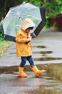 Kid In Yellow Waterproof Cloak, Boots And With Umbrella Playing Outdoors After The Rain