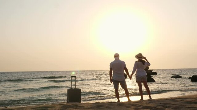 silhouette of couple of lovers. travelers stand on beach, holding hands, against backdrop of sea, sunrise or sunset. there is suitcase nearby. honeymoon, seaside vacation. travel concept.