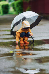 Kid in yellow waterproof cloak, boots and with umbrella playing outdoors after the rain