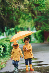 Two kids with umbrella in yellow waterproof cloaks and boots playing outdoors after the rain...