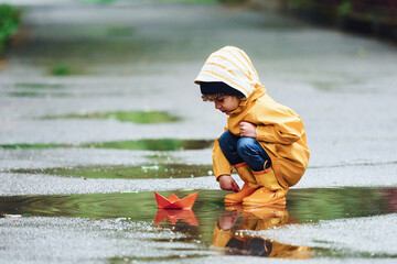 Kid in yellow waterproof cloak and boots playing with paper handmade boat toy outdoors after the rain