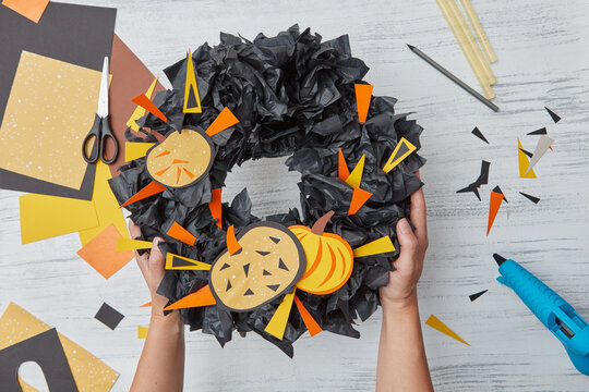 Handmade holiday decoration for interior - Halloween wreath from black paper and colorful pumpkins and applications in a girls hand above wooden table. Flat lay
