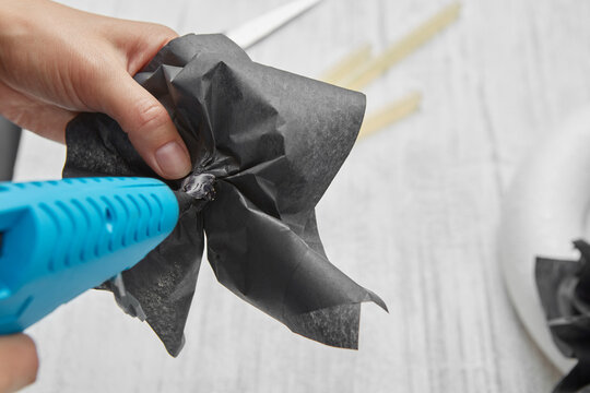Female hands make a black flower from paper with an electric glue gun over a gray wooden table.