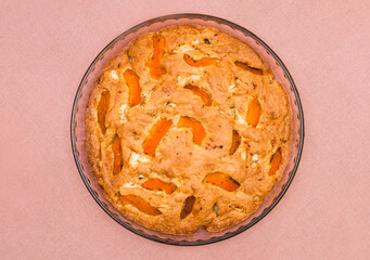 A large, round baked apple and apricot pie stands in a glass form on a table with a pink tablecloth. Delicious, fresh dessert.