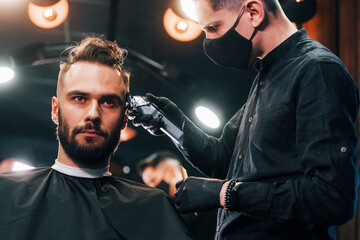 Front view of young bearded man that sitting and getting haircut in barber shop by guy in black...