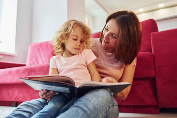 Reading big book. Young mother with her little daughter in casual clothes together indoors at home