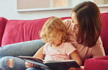 Fototapeta na wymiar Reading big book. Young mother with her little daughter in casual clothes together indoors at home