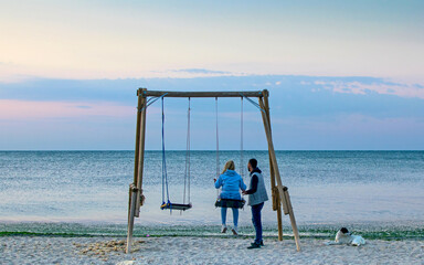 A couple in love on the beach, in the swing. They admire the sunrise. Next to her is a stray puppy. Rear view, no face.