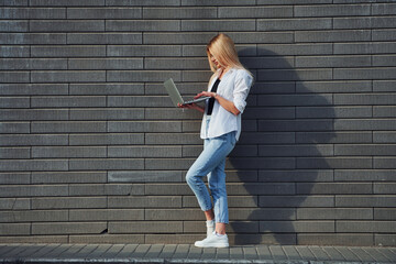 Using laptop. Beautiful blonde in casual clothes is outdoors at sunny daytime