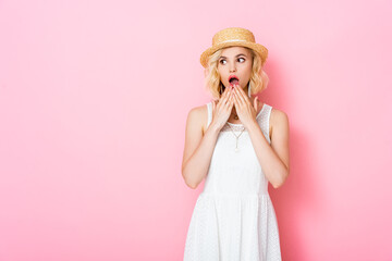 shocked young woman in straw hat covering mouth and looking away on pink