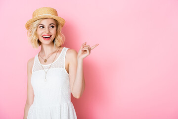 excited woman in straw hat pointing with finger while looking away on pink