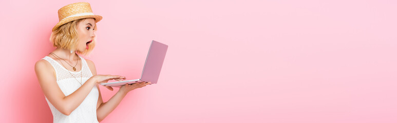 panoramic orientation of shocked woman in straw hat looking at laptop on pink