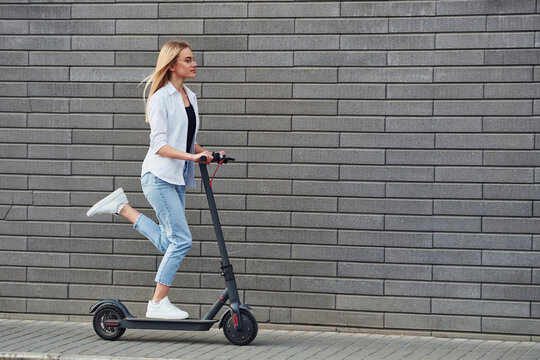Side view of beautiful blonde in casual clothes that riding electric schooter outdoors at sunny daytime