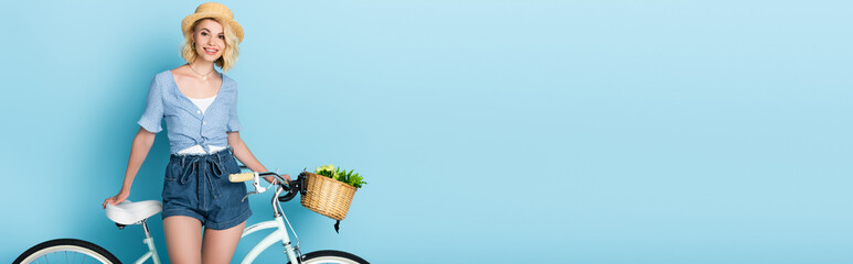 horizontal crop of young woman in straw hat standing near bicycle on blue