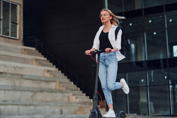 Near business building. Beautiful blonde in casual clothes riding electric schooter outdoors at sunny daytime