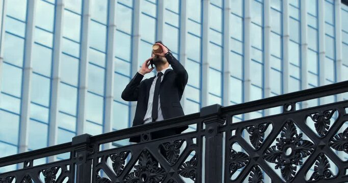 A Man In Office Clothes Went Out To Drink Coffee And Talk On The Phone During Lunch On The Bridge Against The Backdrop Of The Blue Windows Of The Building.