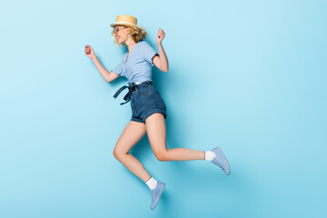side view of young woman in straw hat and sunglasses jumping on blue