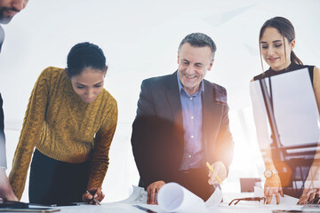 Smiling crew of architects satisfied with accomplished blueprint for project drafting together in office.Mature boss checking work of experienced engineers cooperating with employees in good mood