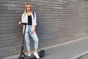 Fototapeta na wymiar Against grey wall. Beautiful blonde in casual clothes riding electric schooter outdoors at sunny daytime