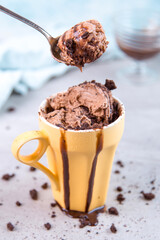 Chocolate brown ice cream in a regular yellow mug and spoon, on a blue background, with topping and crumbs. Unusual serve of dessert, copy space.