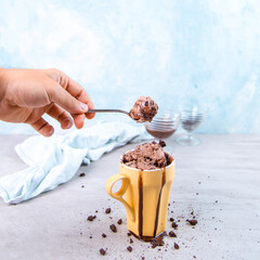 Chocolate brown ice cream in a regular yellow mug and spoon, on a blue background, with topping and crumbs. Unusual serve of dessert, copy space.